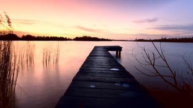Holzsteg führt über ruhiges Wasser, umgeben von Gräsern und einem farbenprächtigen Sonnenuntergang.
