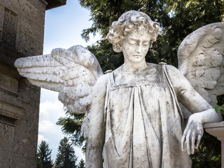 Skulptur eines Engel mit Flügeln, gefaltetem Arm und nachdenklichem Gesicht, vor einer Mauer und grünen Bäumen.
